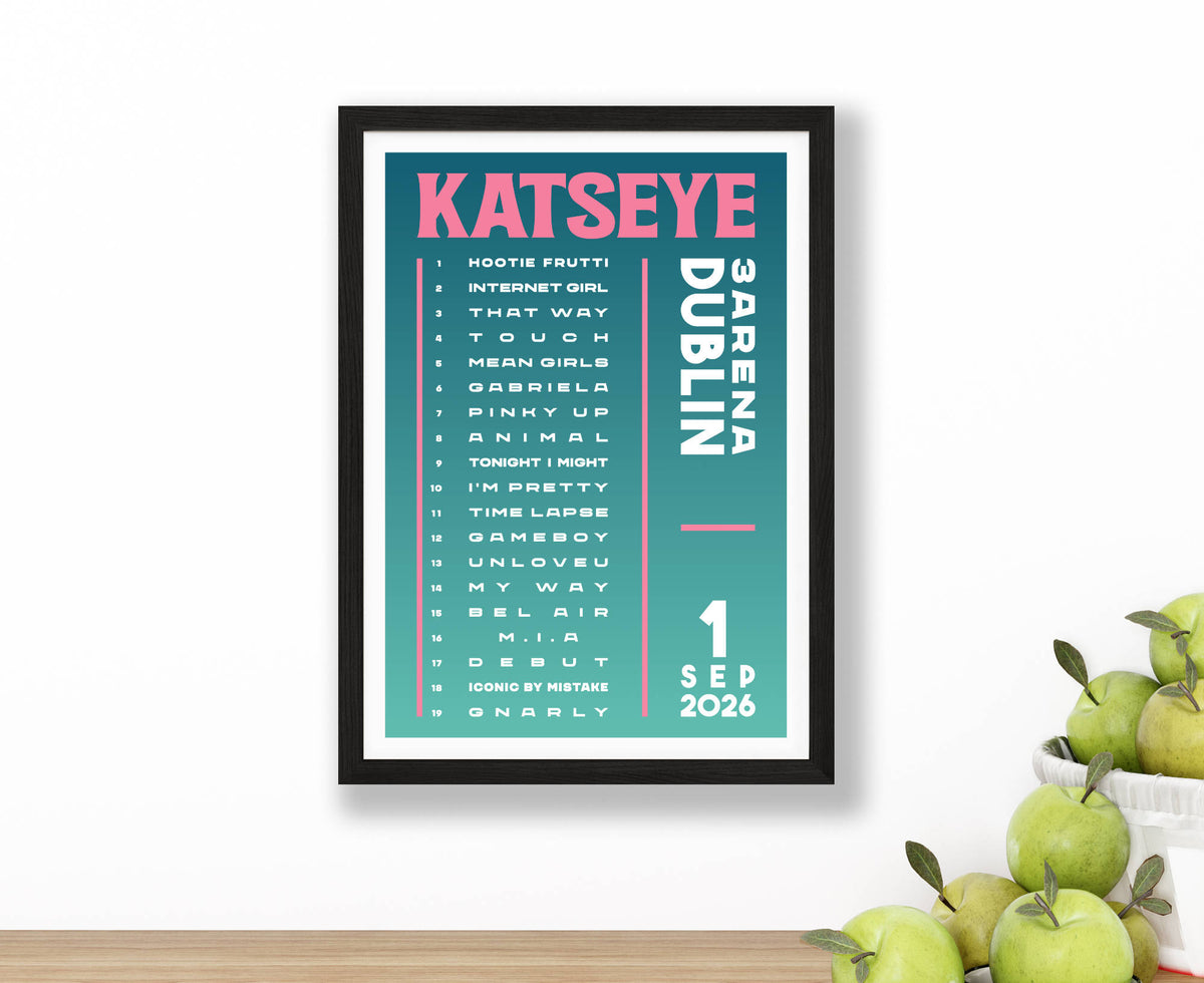 A setlist poster for Katseye at 3Arena Dublin on 1 September 2026, featuring a teal background with pink and white text, displayed on a wooden surface next to green apples against a white wall.