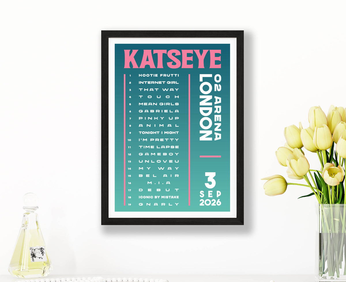 A framed music poster for Katseye's London concert on 3 Sep 2026, featuring a teal background with pink and white text, styled on a white wall next to yellow tulips in a vase.