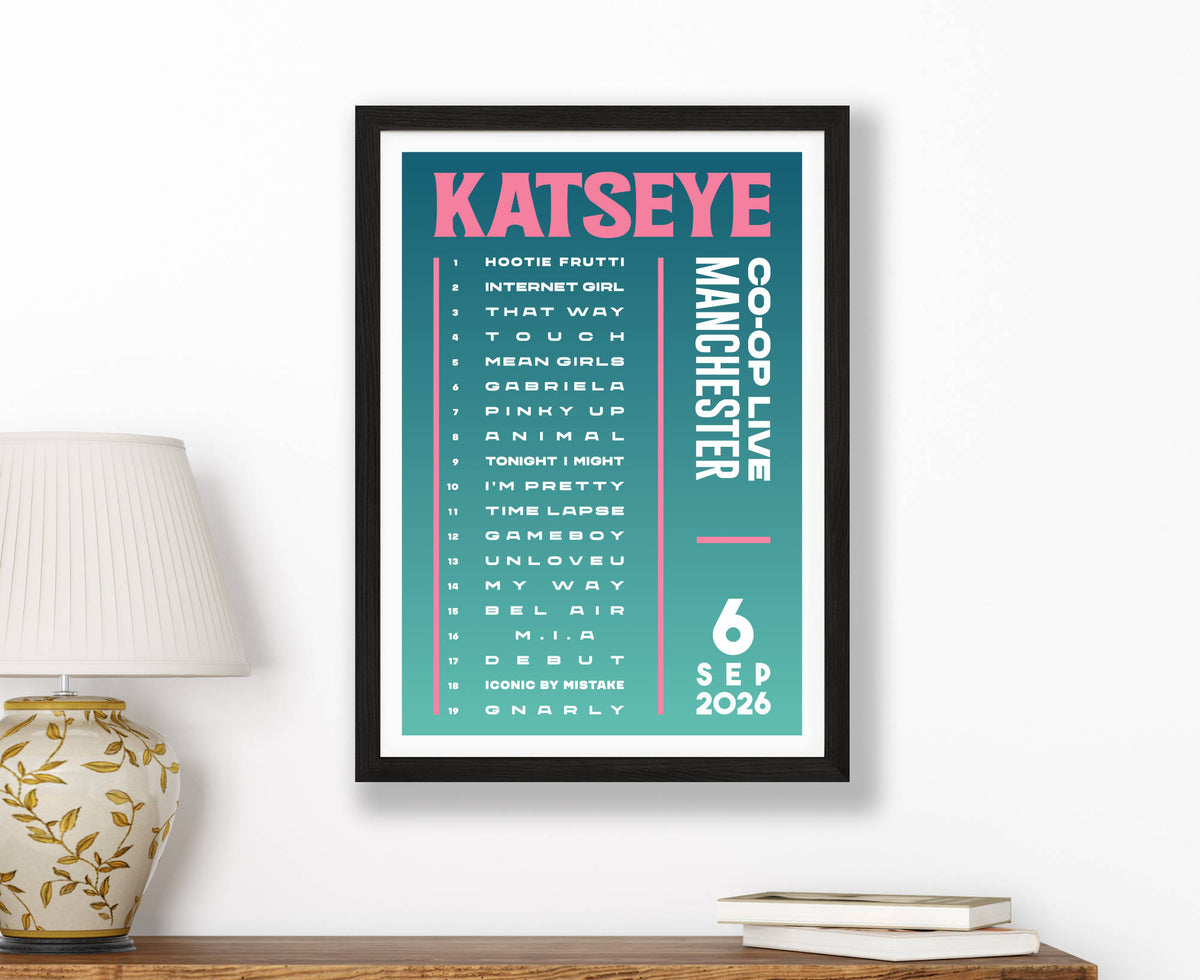 A framed music commemorative print for Katseye's Co-Op Live Manchester show on a white wall above a wooden shelf with a lamp and books. The print features a teal background with pink and white text.
